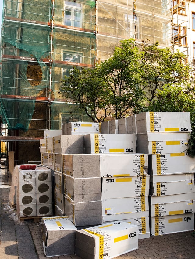 Gestapelte Dämmplatten vor einem eingerüsteten Altbau mit grünem Gerüstnetz