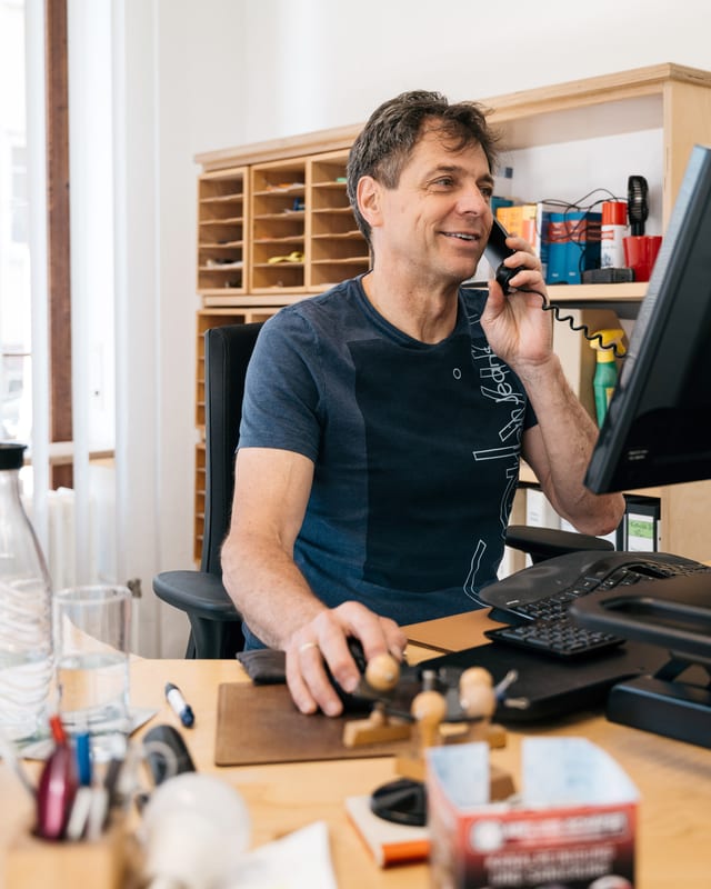 Ralf Schäfer telefoniert an seinem Schreibtisch, vor ihm der Monitor, hinter ihm ein Fächerregal